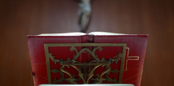 Libro rojo con detalles dorados abierto sobre un soporte, con una figura de un crucifijo desenfocada en el fondo.